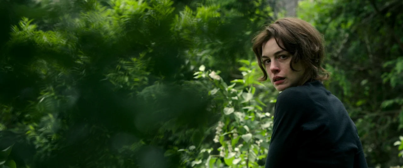 Mother’s Instinct (2024) movie still: medium — A woman stands within a dense thicket of green foliage and leafy branches. The subject turns her head…