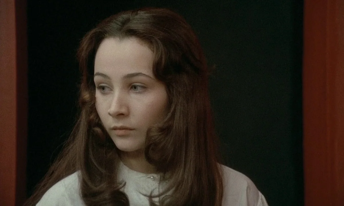 Immoral Tales (1974) movie still: closeup — A woman stands before a dark surface flanked by reddish-brown vertical wooden framing. She gazes…