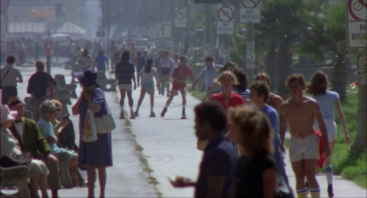 Xanadu (1980) movie still: wide — A sunny public boardwalk is crowded with people walking and skating near palm trees. Numerous metal signage poles…