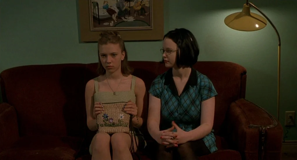 Ghost World (2001) movie still: medium — Two teenage girls sit on a dark couch against a plain teal wall. A framed illustration hangs above them and…