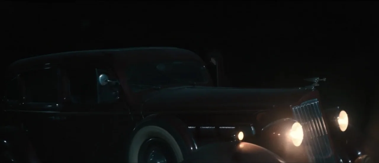Fear Below (2025) movie still: medium — A dark 1930s vintage sedan occupies the frame in a pitch black setting. Its bright round headlights pierce…