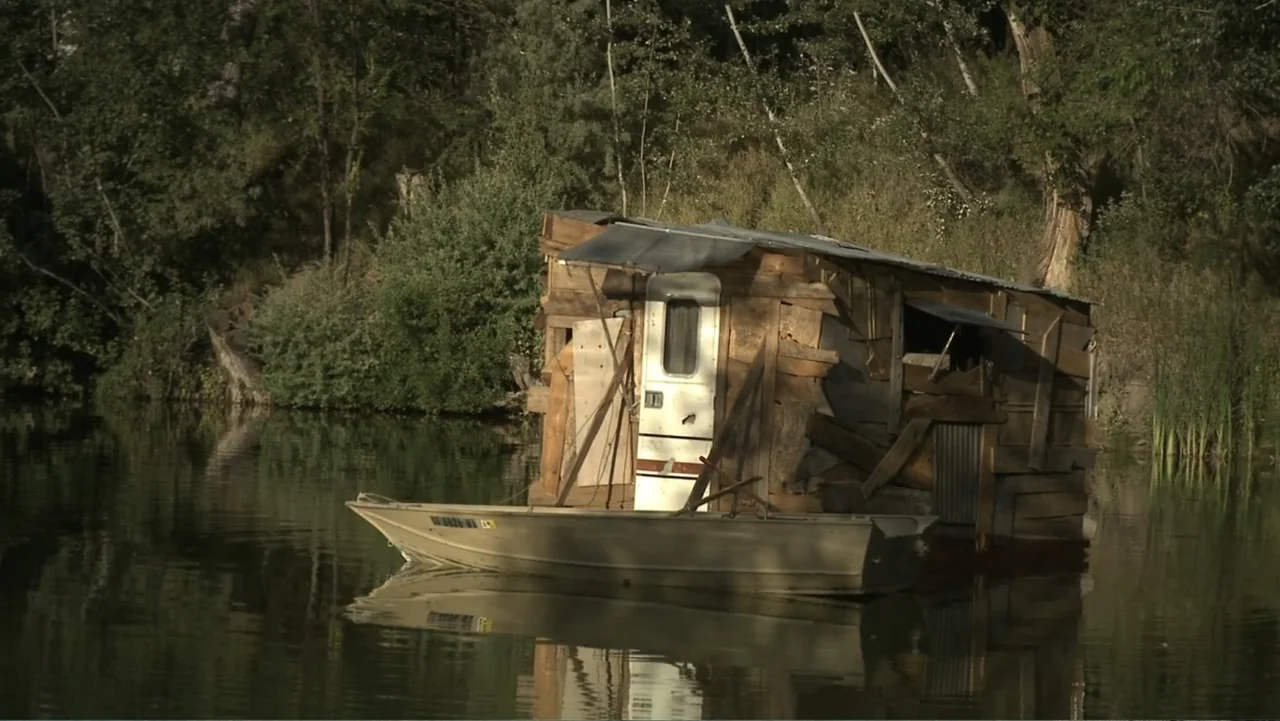 Boathouse Detectives (2010) movie still: medium — A makeshift wooden houseboat structure sits on a calm body of water. A green metal skiff is…