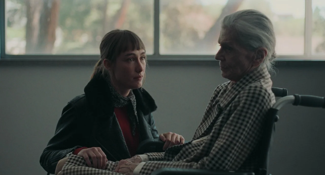 La Abuela (2021) movie still: medium — A spare, neutral room with a large window in the background. A young woman kneels beside an elderly woman…