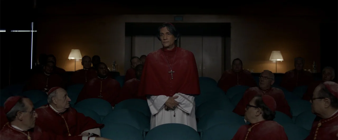 Conclave (2024) movie still: medium — A dimly lit auditorium features rows of blue seats occupied by individuals in ecclesiastical vestments. A man…