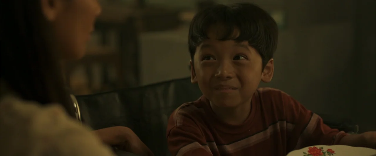 The Unholy (2022) movie still: medium — An interior dining area with dim, low-key lighting and muted green and brown tones. A young boy smiles while…