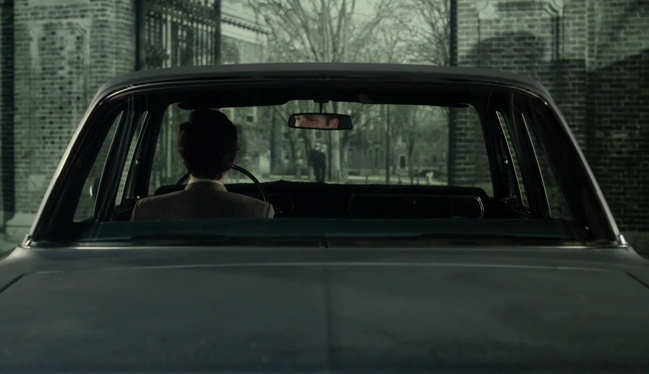 Experimenter (2015) movie still: wide — A dark car sits on a paved driveway in front of a brick building. The driver is viewed from behind as his…