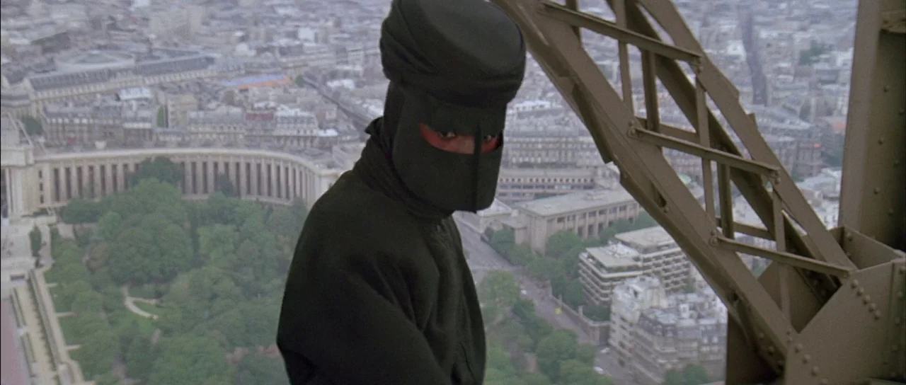 A View to a Kill (1985) movie still: medium — A figure stands on the iron structure of the Eiffel Tower overlooking a city. The subject wears a dark…