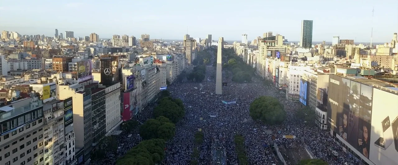 I Choose to Believe (2023) movie still: establishing — A wide aerial view shows a dense city avenue filled with a massive crowd. A large stone…