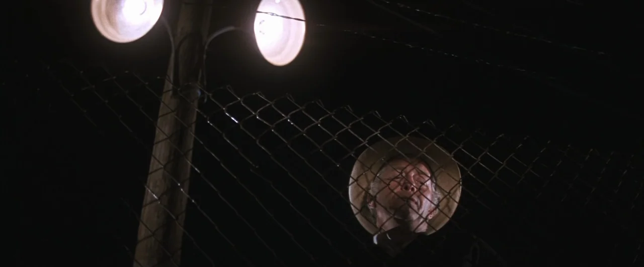 Cool Hand Luke (1967) movie still: medium — An older man stands behind a metal chain link fence at night. He is illuminated from above by two bright…