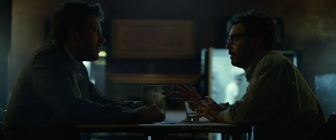 Gone Girl (2014) movie still: medium — A dark, dimly lit diner booth interior at night with wooden table surface. Two men sit in profile across from…
