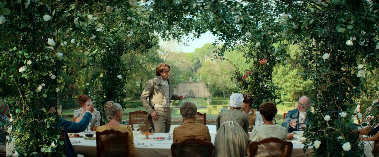 The Count of Monte-Cristo (2024) movie still: wide — A group of people sits at an outdoor table under a rose-covered archway. A man stands at the…