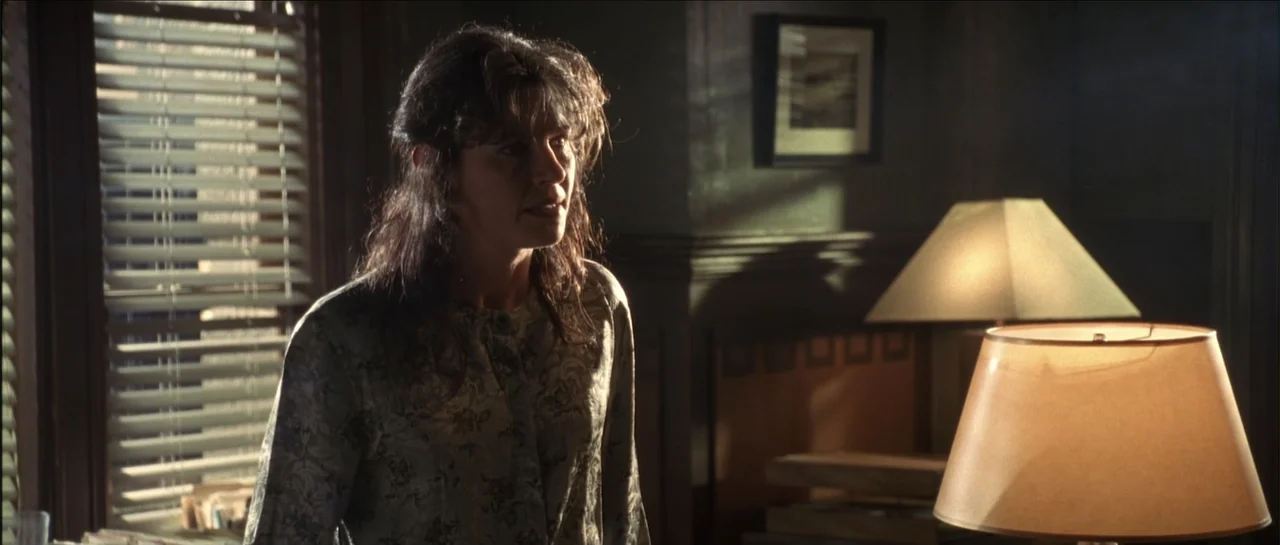 Gang Related (1997) movie still: medium — An interior office with dark wood paneling and window blinds filtering daylight. A woman stands in the…