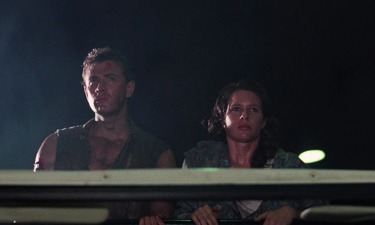 Demons (1985) movie still: medium — A man and woman stand in the back of a moving vehicle at night. The surrounding environment is obscured by…