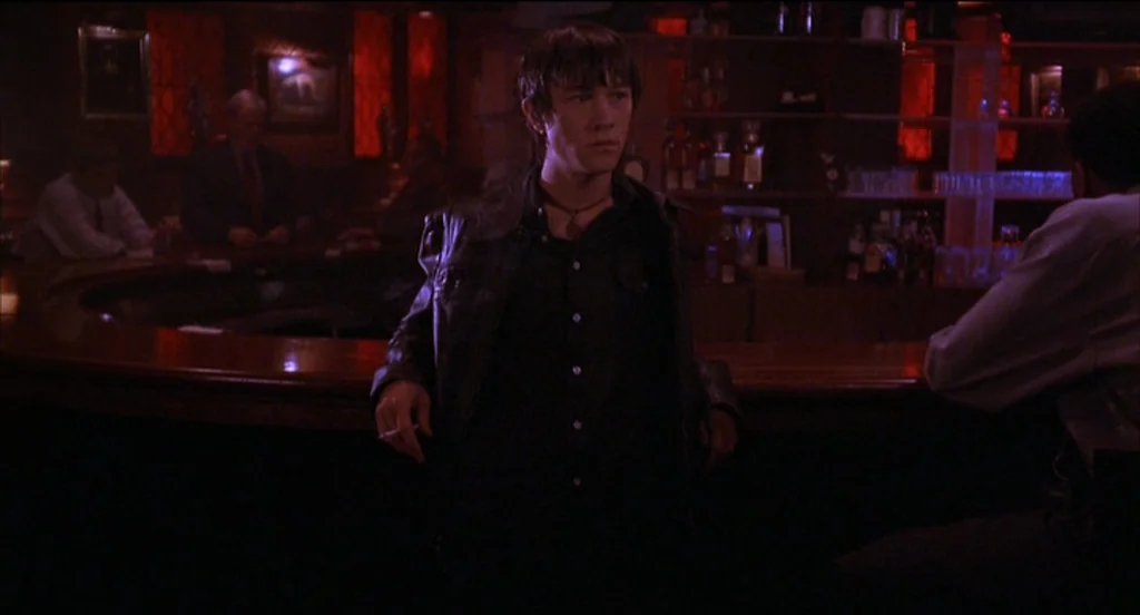 Mysterious Skin (2004) movie still: medium — A dimly lit bar with red-toned wood paneling and shelves filled with liquor bottles. A young man with…