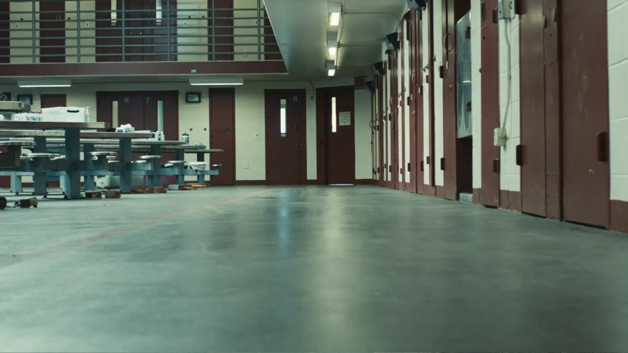 Camp X-Ray (2014) movie still: establishing — A prison cell block features a polished concrete floor and rows of red steel doors. Empty tables are…