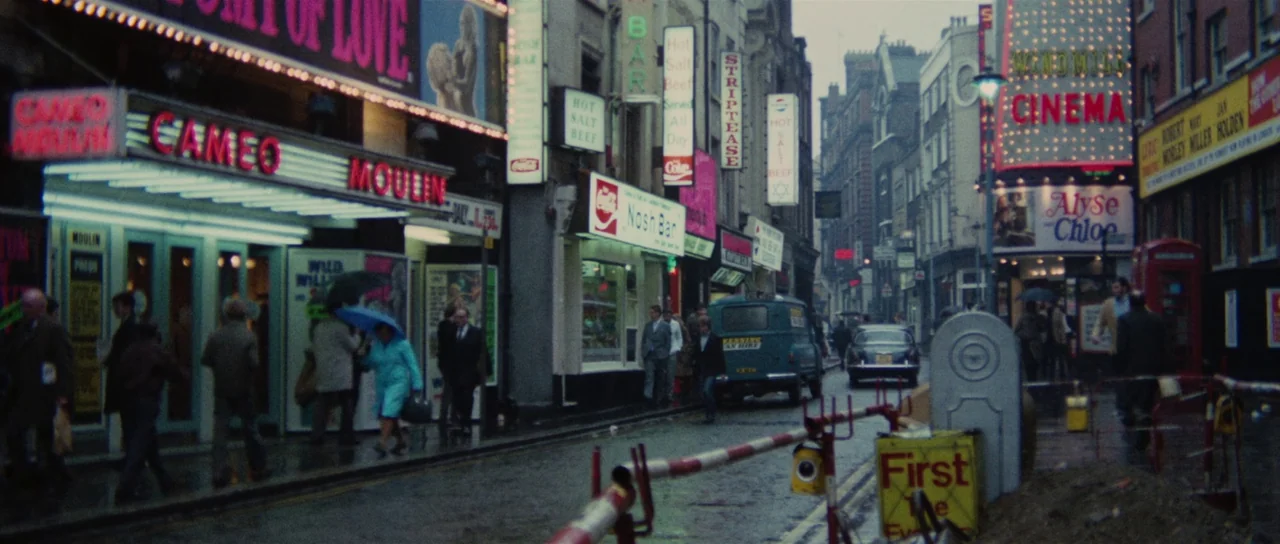 Death Walks On High Heels (1971) movie still: wide — An urban street in 1970s London featuring neon cinema signs and rainy pavement. Pedestrians…