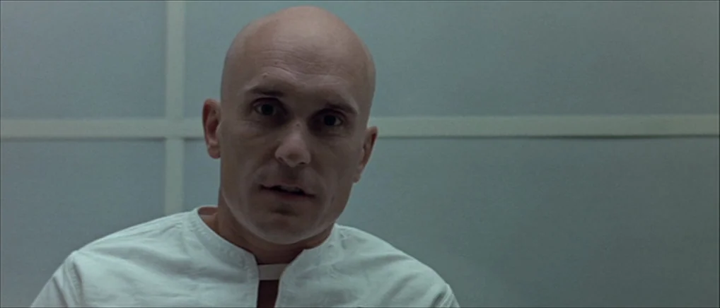 THX 1138 (1971) movie still: closeup — A sterile medical room features a grid-patterned backlit wall with translucent panels. A bald man wearing a…