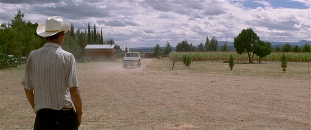 Silent Light (2007) movie still: medium — A rural farm landscape features a dirt driveway and a distant field of crops. A man in a cowboy hat stands…