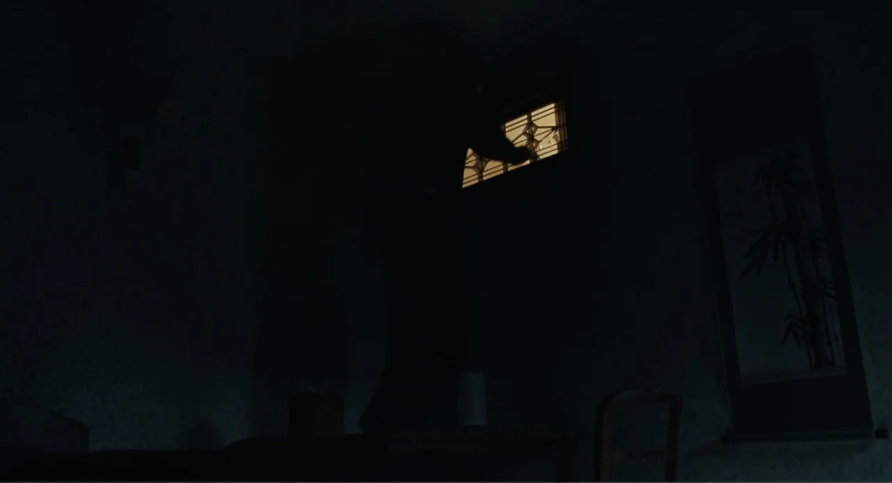 Winter in Sokcho (2025) movie still: medium — A dark, dimly lit room contains a person standing on a wooden table. The subject reaches toward a…
