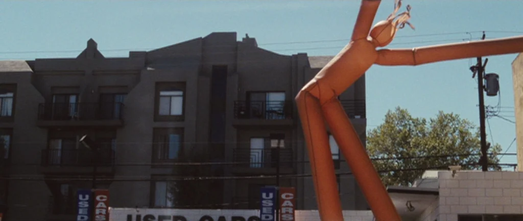 Greenberg (2010) movie still: wide — A used car lot features an orange inflatable tube man swaying in the air. In the background, a multi-story…