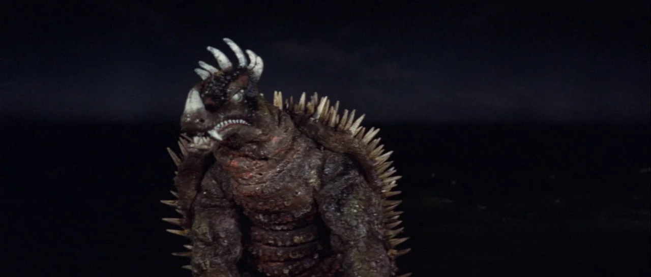 Godzilla Vs Gigan (1972) movie still: medium — A bipedal, spike-covered reptilian monster stands in a dark, empty, and undefined landscape. The…