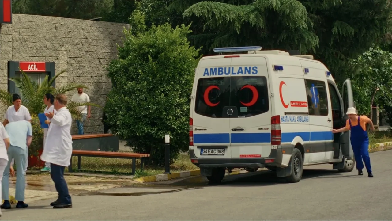 Paranormal Cuma (2024) movie still: wide — A hospital emergency department exterior with stone walls and a parked white ambulance van. Medical staff…