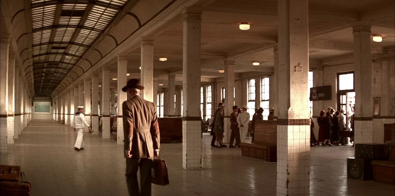 The Last Emperor (1987) movie still: wide — A spacious, tiled passenger hall with rows of support columns and wooden benches. A man in a suit walks…