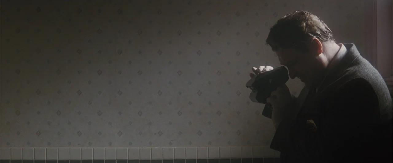 Fallen (2004) movie still: medium — A dimly lit room with patterned wallpaper above a tiled wainscoting. A man in a dark blazer uses a vintage…
