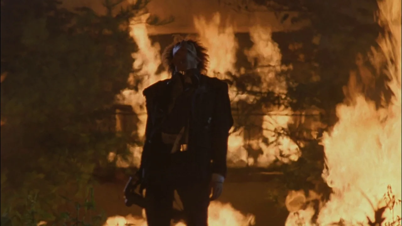 Battle Royale (2000) movie still: fullbody — A figure stands in a wooded area surrounded by large, uncontrolled roaring flames. The individual looks…