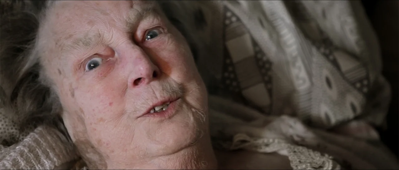 Garnet’s Gold (2014) movie still: closeup — An interior bedroom setting featuring an elderly woman resting against soft white patterned pillows. The…