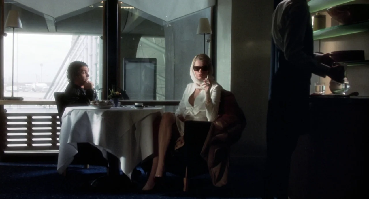 Femme Fatale (2002) movie still: wide — An airport lounge with a white-clothed table set against a large window view. A woman sits at the table…