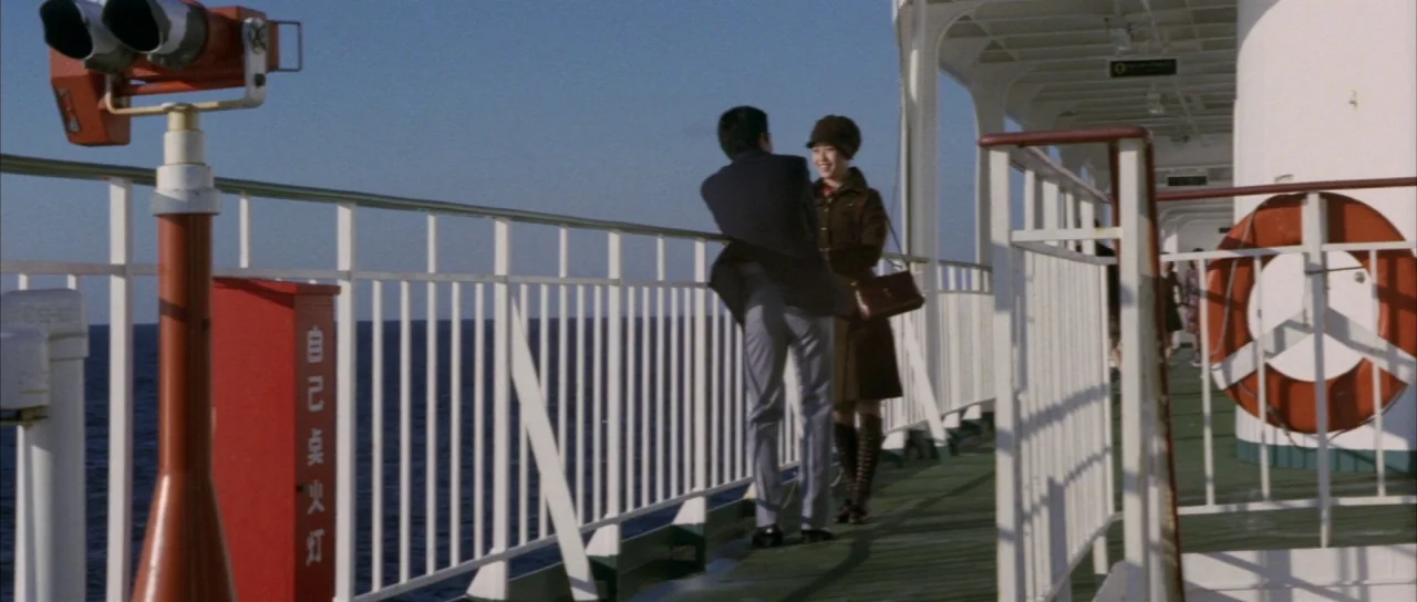 Godzilla Vs Mechagodzilla (1974) movie still: wide — A ferry deck with a white railing overlooks a dark blue ocean expanse. A man and a woman stand…
