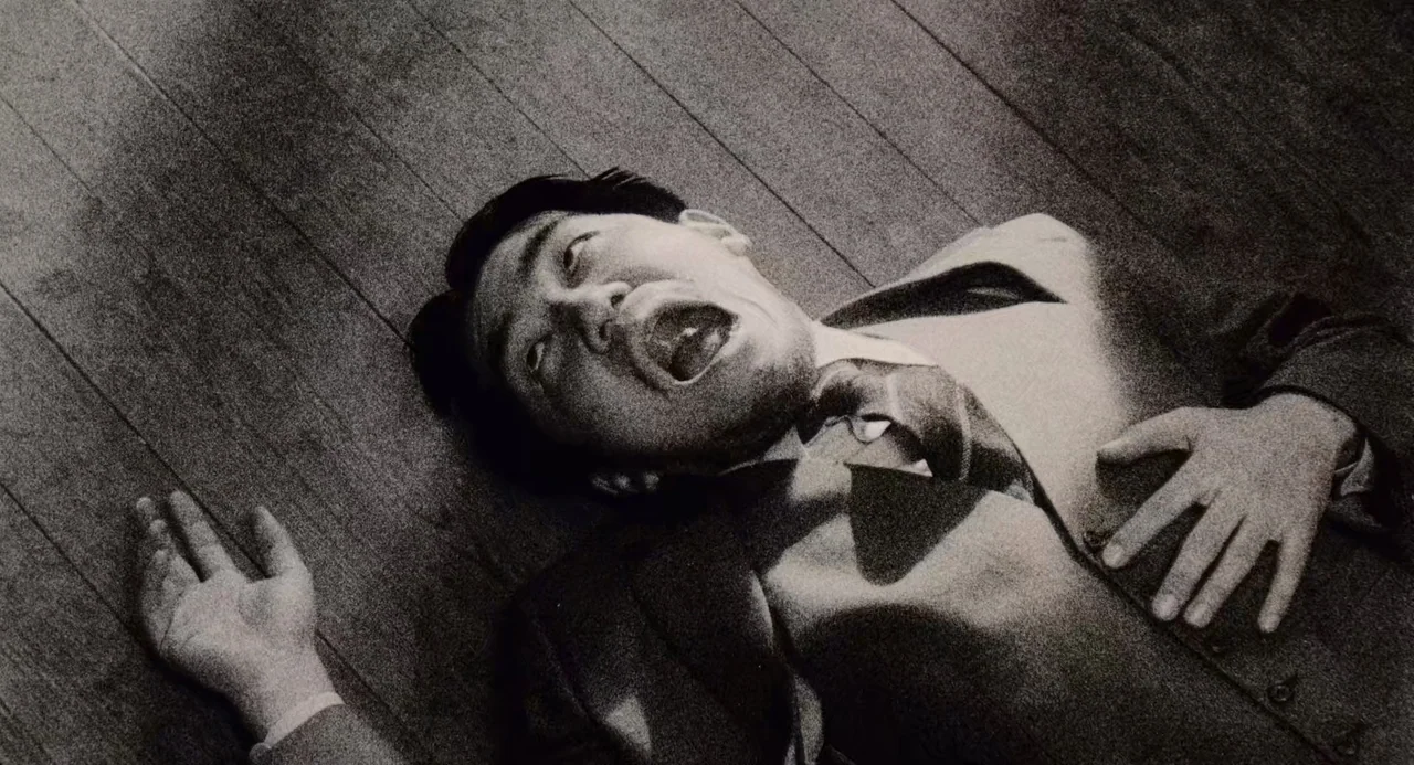 Ringu (1998) movie still: medium — A man lies prone on a wooden floor in a dimly lit room. He is dressed in a suit with his head tilted back.