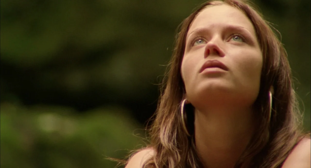 My Summer of Love movie still: closeup — A woman stands in a densely vegetated wooded area with blurred foliage behind her. She looks upward with an…