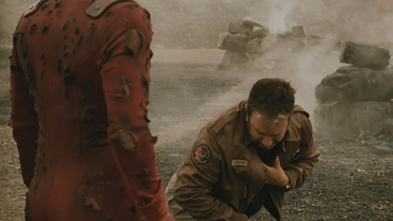 Zoom (2006) movie still: medium — A desolate and rocky landscape filled with thin smoke and scattered debris. A man in a brown jacket kneels beside…