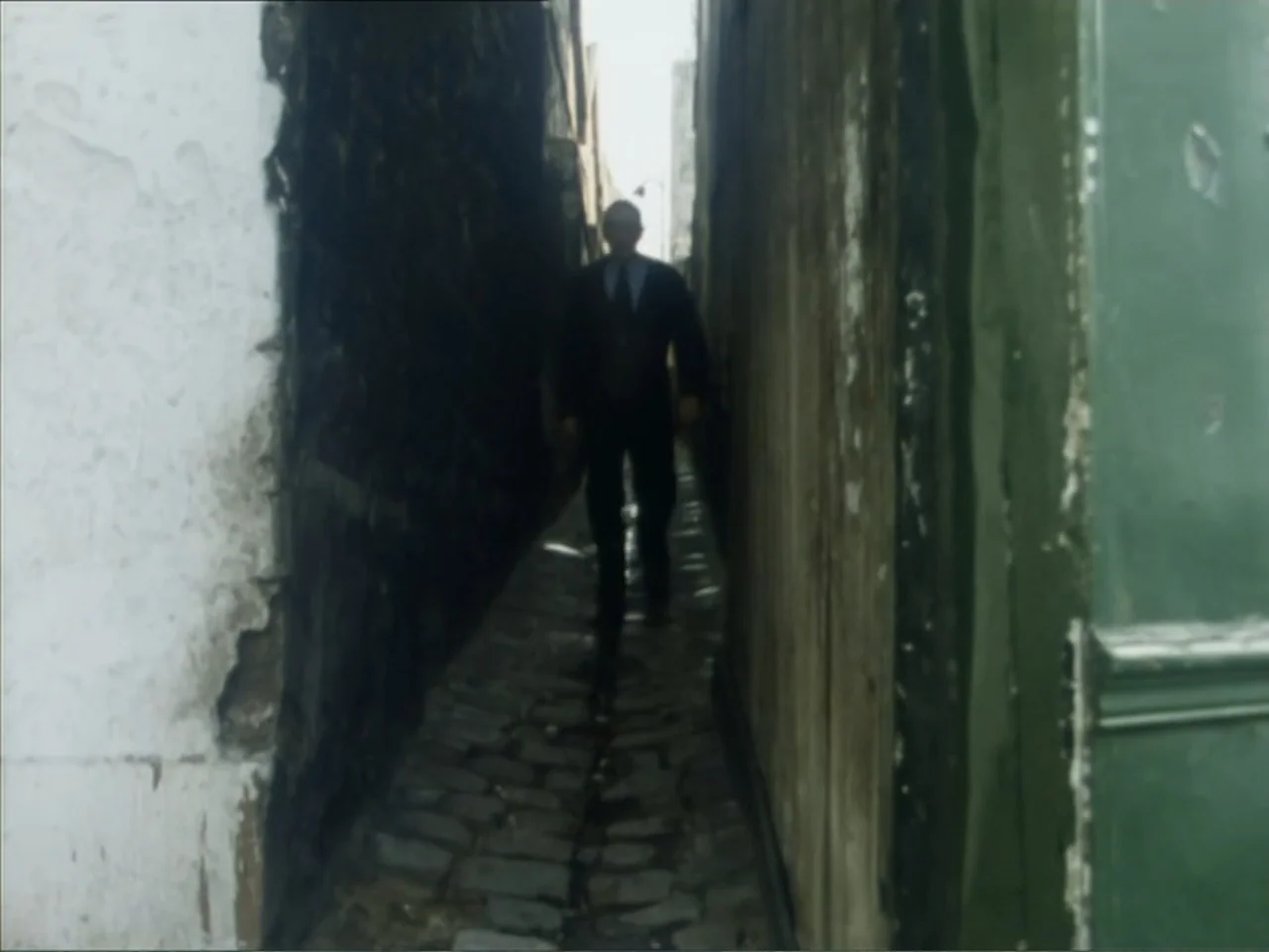 A Dessert for Constance (1981) movie still: fullbody — A narrow cobblestone alleyway is framed by weathered stone and painted wooden walls. A man in…