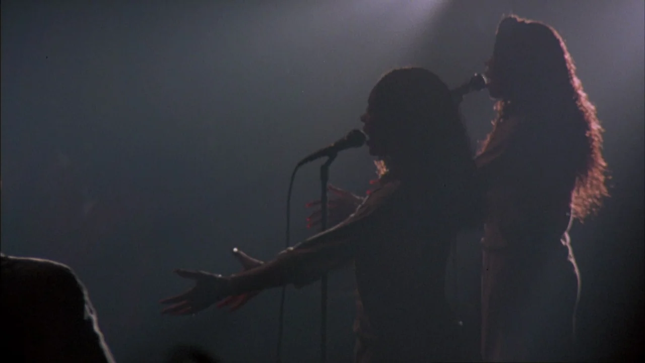 Stop Making Sense (1984) movie still: medium — Two women stand on a dark stage with spotlights highlighting their silhouettes. They are singing into…
