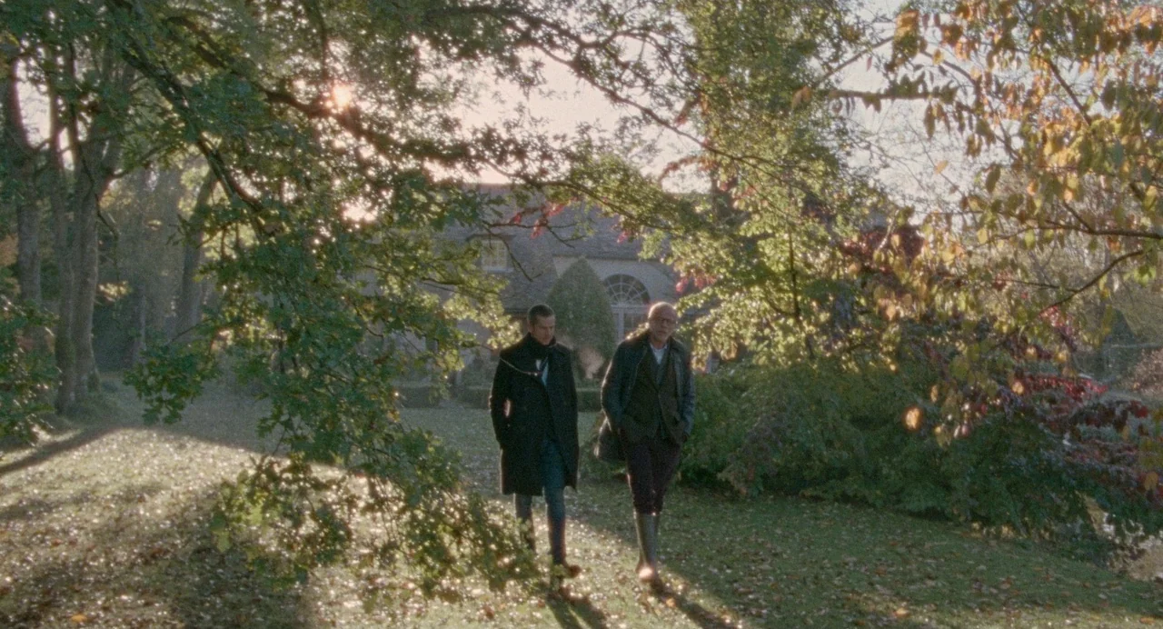 Non-Fiction (2018) movie still: wide — A stone manor house sits in the background of a lush, tree-lined property. Two men walk together across the…