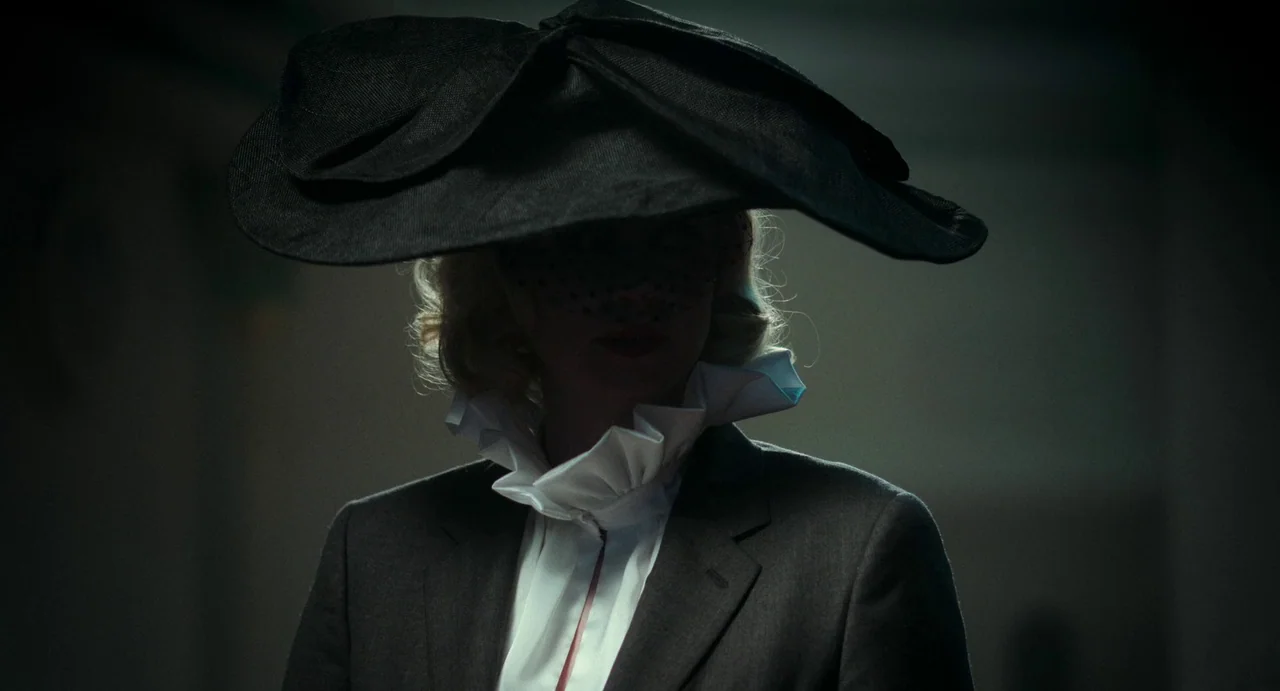 Flux Gourmet (2022) movie still: closeup — A dimly lit interior space contains a subject wearing a large brimmed hat. The woman wears a dark blazer…