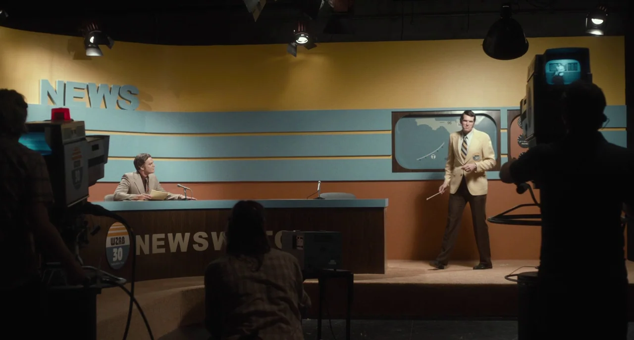 Christine (2016) movie still: wide — A 1970s television news studio set features yellow and blue walls with wooden desk panels. One man sits behind…