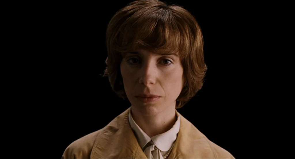 Submarine movie still: closeup — A woman stands centered against a completely black, non-descript background. She looks directly toward the camera…