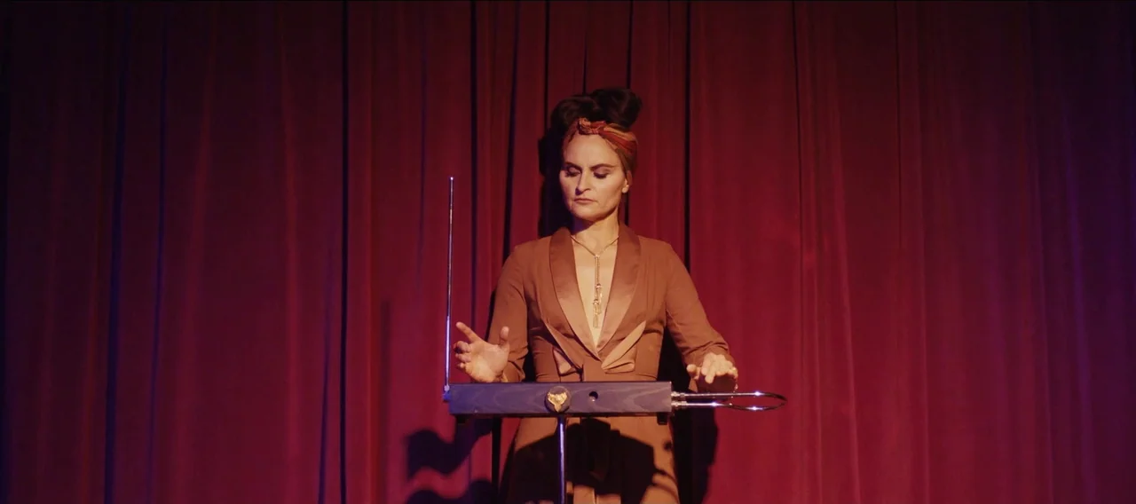 Fuzzy Head (2023) movie still: medium — A woman stands before a deep red curtain on a dimly lit stage. She operates a theremin by moving her hands…