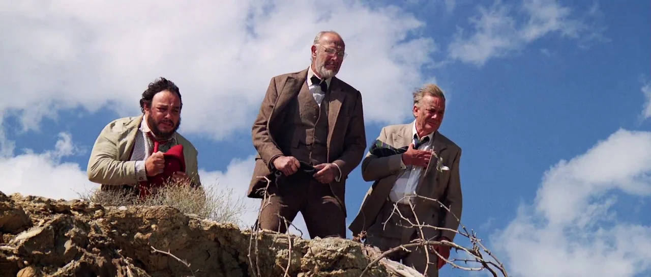 Indiana Jones and the Last Crusade (1989) movie still: fullbody — Three men in suits stand atop a dry, rocky desert ridge under a blue sky. The men…