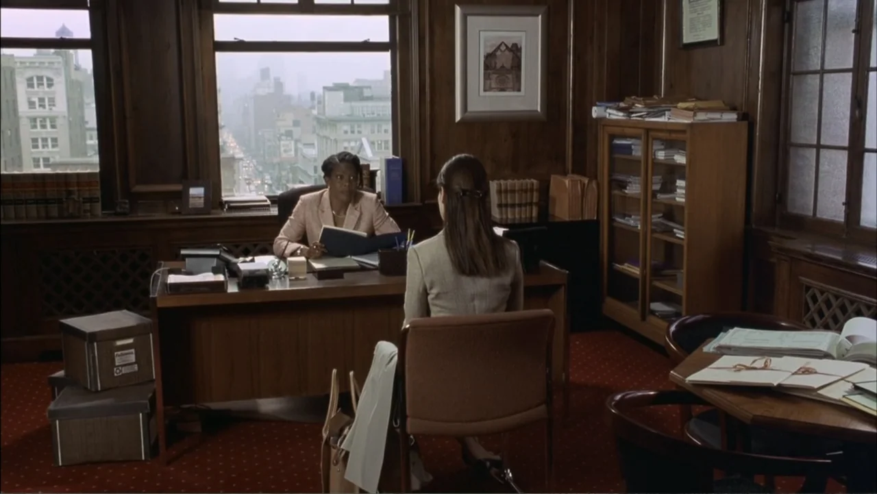 Two Weeks Notice (2002) movie still: medium — A wood-paneled law office features a large window overlooking a city street. A woman sits behind the…