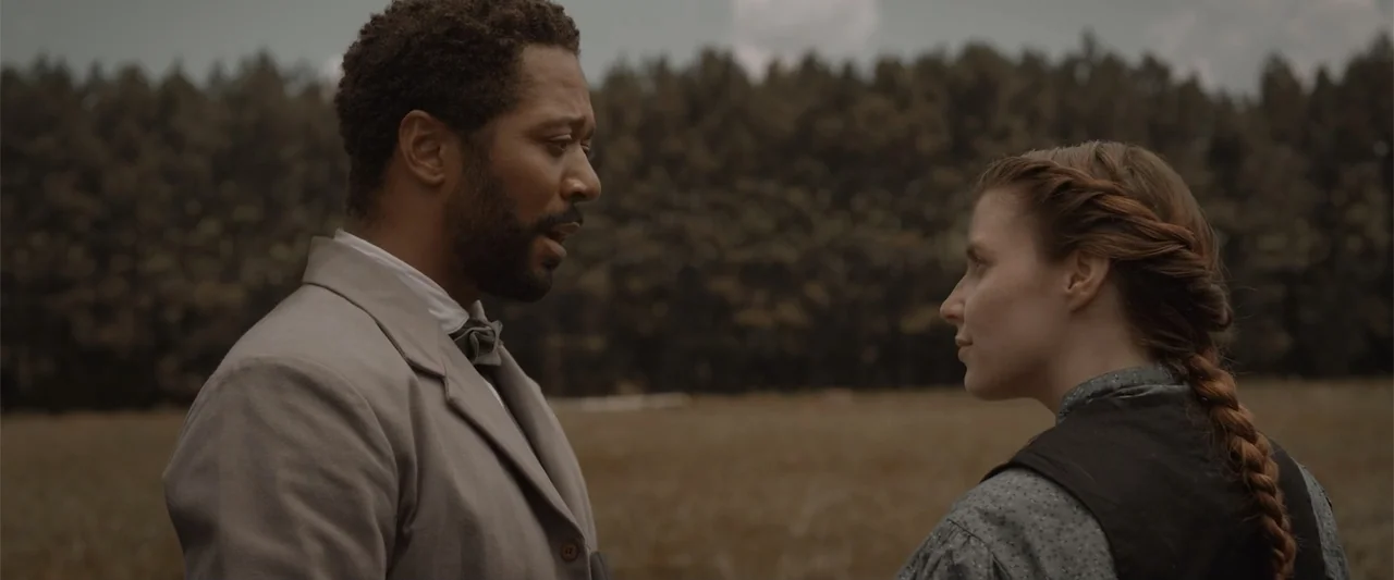 Ghosts of the Ozarks (2022) movie still: medium — An open field with a dense treeline in the background under an overcast sky. A man and woman in…