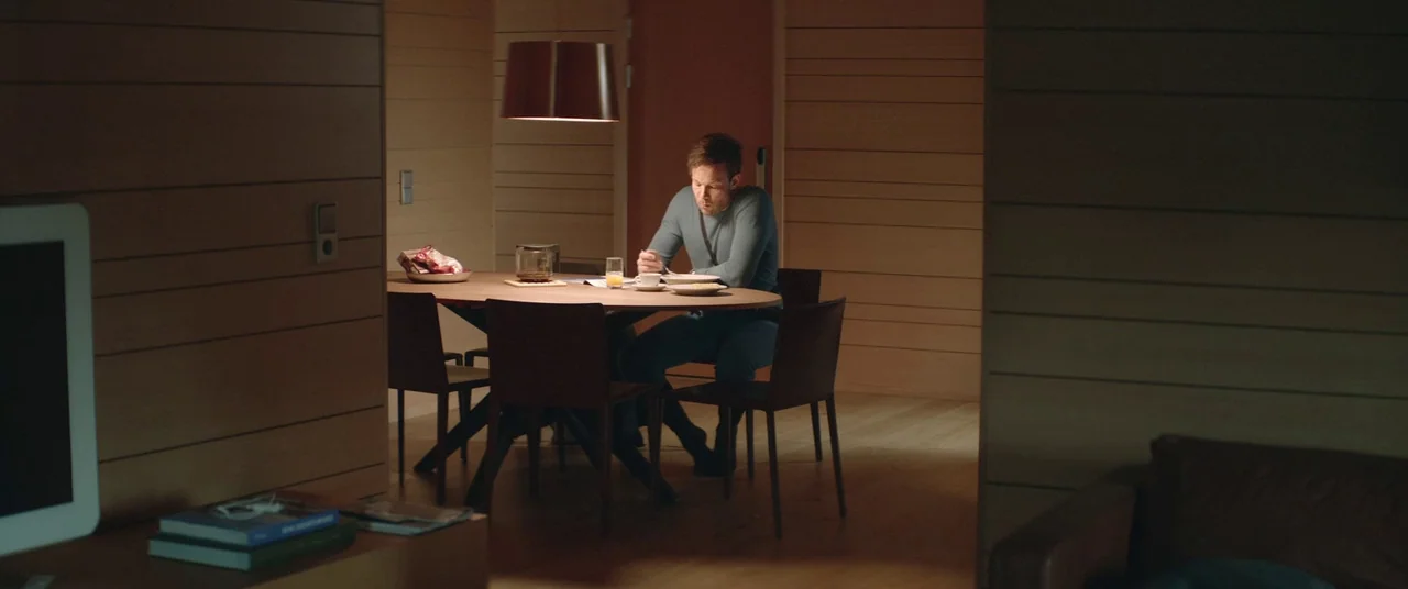 Force Majeure (2014) movie still: wide — A modern, minimalist space with horizontal wooden wall panels and a simple oval table. A man eats alone…