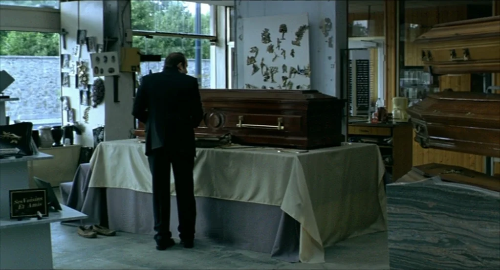 On War (2008) movie still: medium — A funeral home showroom interior features several wooden coffins displayed on tables and stands. A man in a dark…