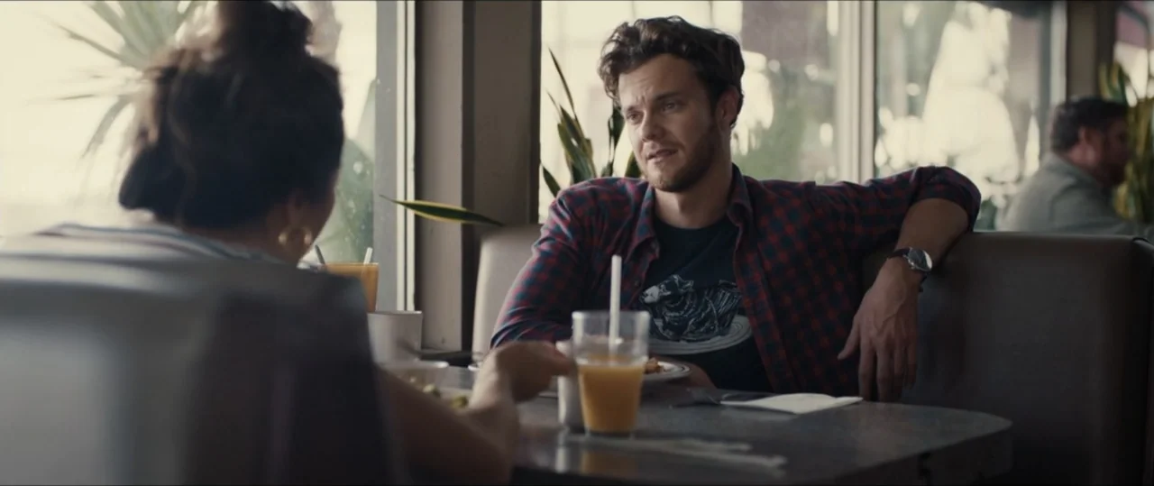 Plus One (2019) movie still: medium — A diner booth interior features dark padded seats and a gray laminate table surface. A young man sits opposite…