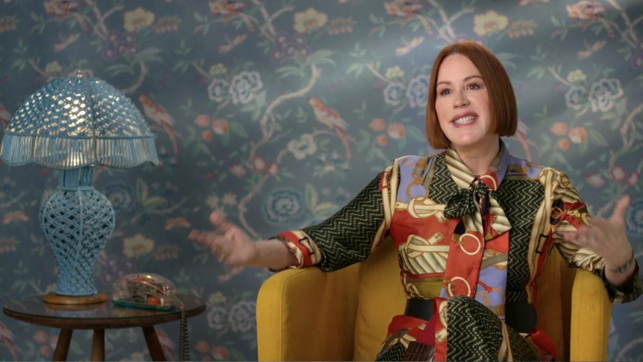 Judy Blume Forever (2023) movie still: medium — A woman sits in a yellow armchair in front of a blue floral wallpaper. She gestures with both hands…