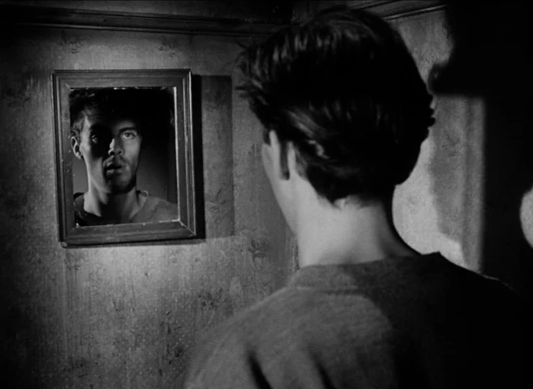 The Blue Lamp (1950) movie still: medium — A dimly lit room features textured wallpaper and a small square wooden mirror. A man stands facing his…